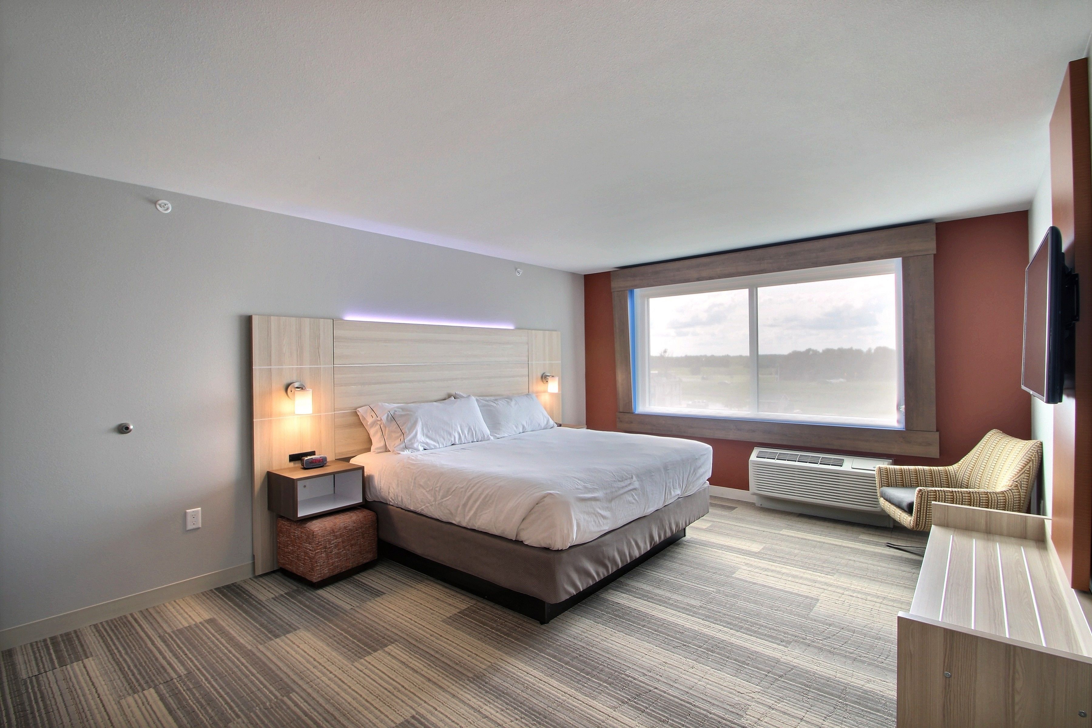 Una habitación de hotel moderna con una cama grande, una mesita de noche, una silla, un ventanal con vistas y un equipo de aire acondicionado integrado.