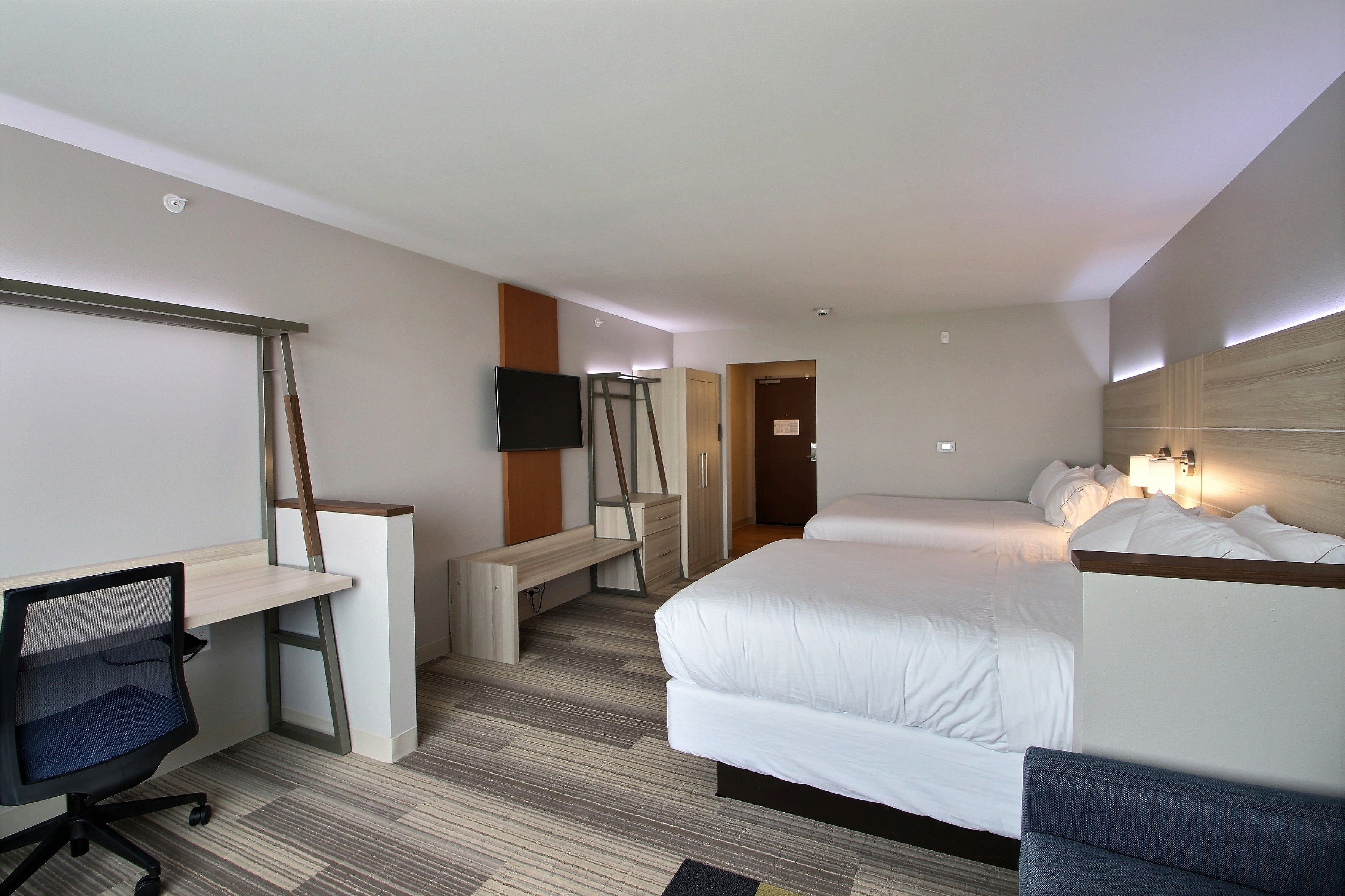 A hotel room with a bed, a desk, a chair, a TV, and a bench.