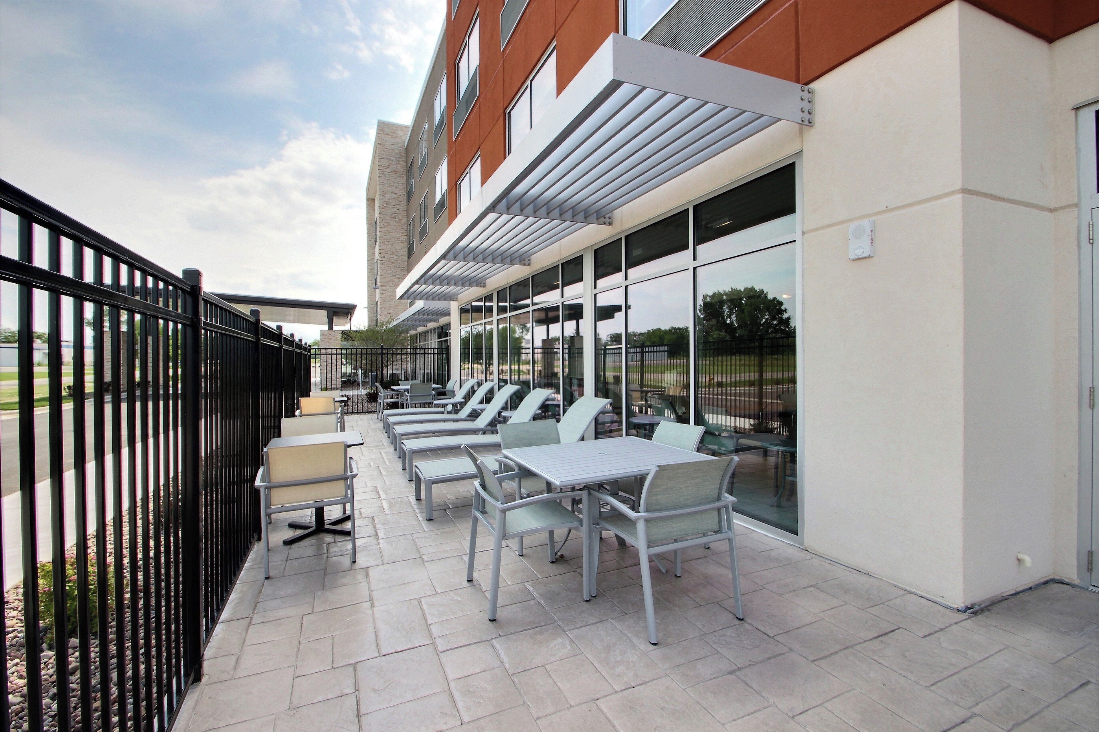 An outdoor patio with tables and chairs, a black metal fence, and a building with large windows.