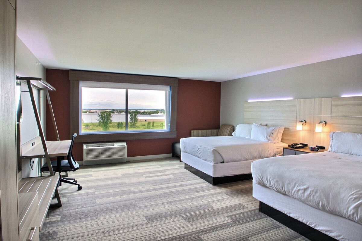 Een hotelkamer met twee bedden, een bureau, een stoel, een groot raam en uitzicht op de omgeving.