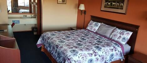 A hotel room with a bed, a desk, a mirror, a sink, and a framed picture on the wall.