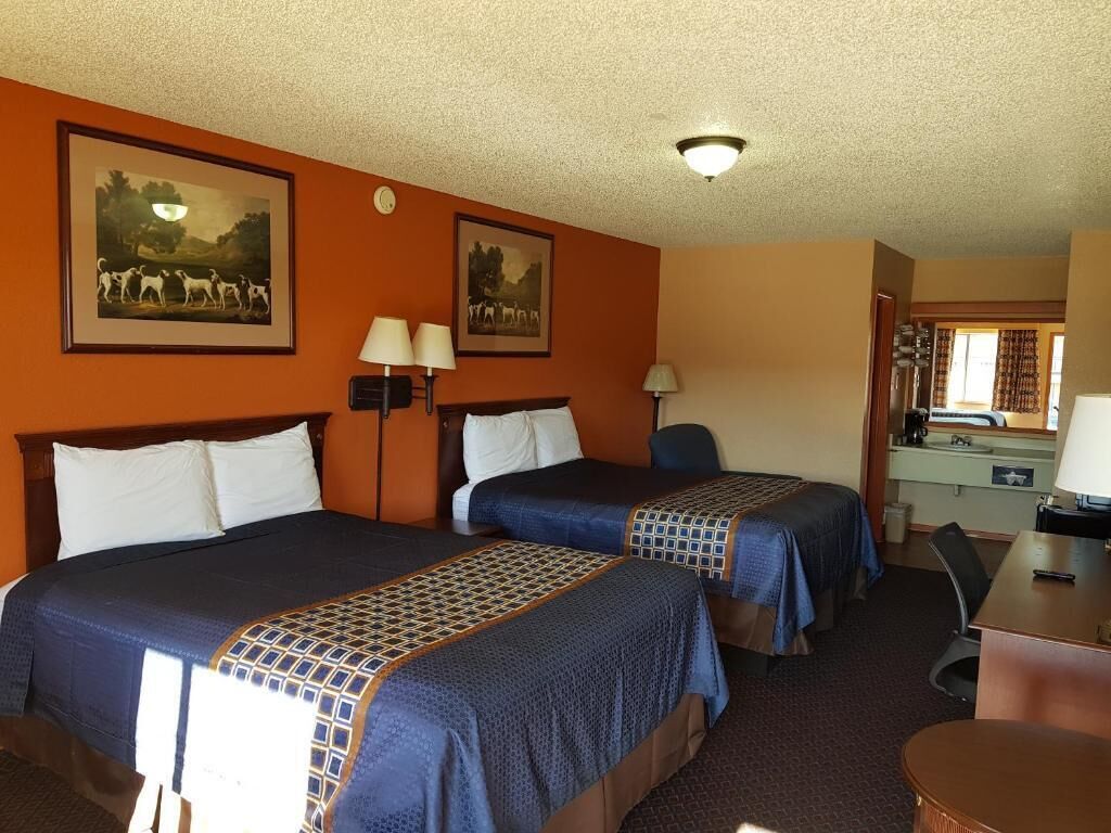 Habitación de hotel con dos camas, un escritorio y una silla. Hay dos lámparas en el escritorio y un cuadro en la pared.