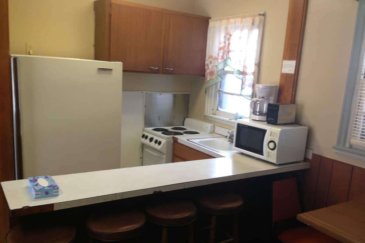 A compact kitchen with a refrigerator, stove, sink, and a microwave on the counter. There are stools under the counter and a window with curtains.
