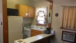 A compact kitchen with wooden cabinets, a stove, a microwave, and a small window with curtains. - Jamestown Motel (Jamestown)