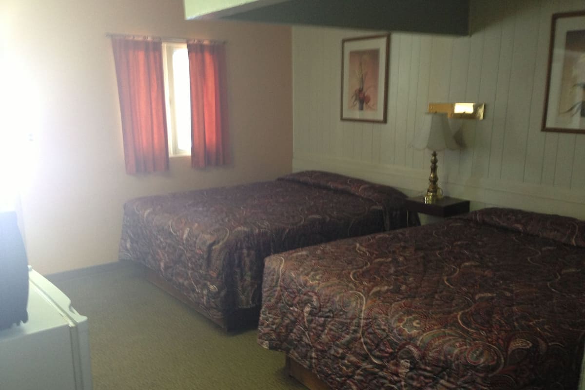 A room with two beds, a window with red curtains, a framed picture on the wall, and a bedside table with a lamp.