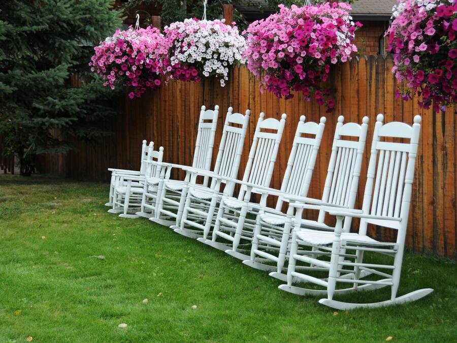 Una hilera de mecedoras blancas alineadas junto a una valla de madera con cestas de flores colgantes.