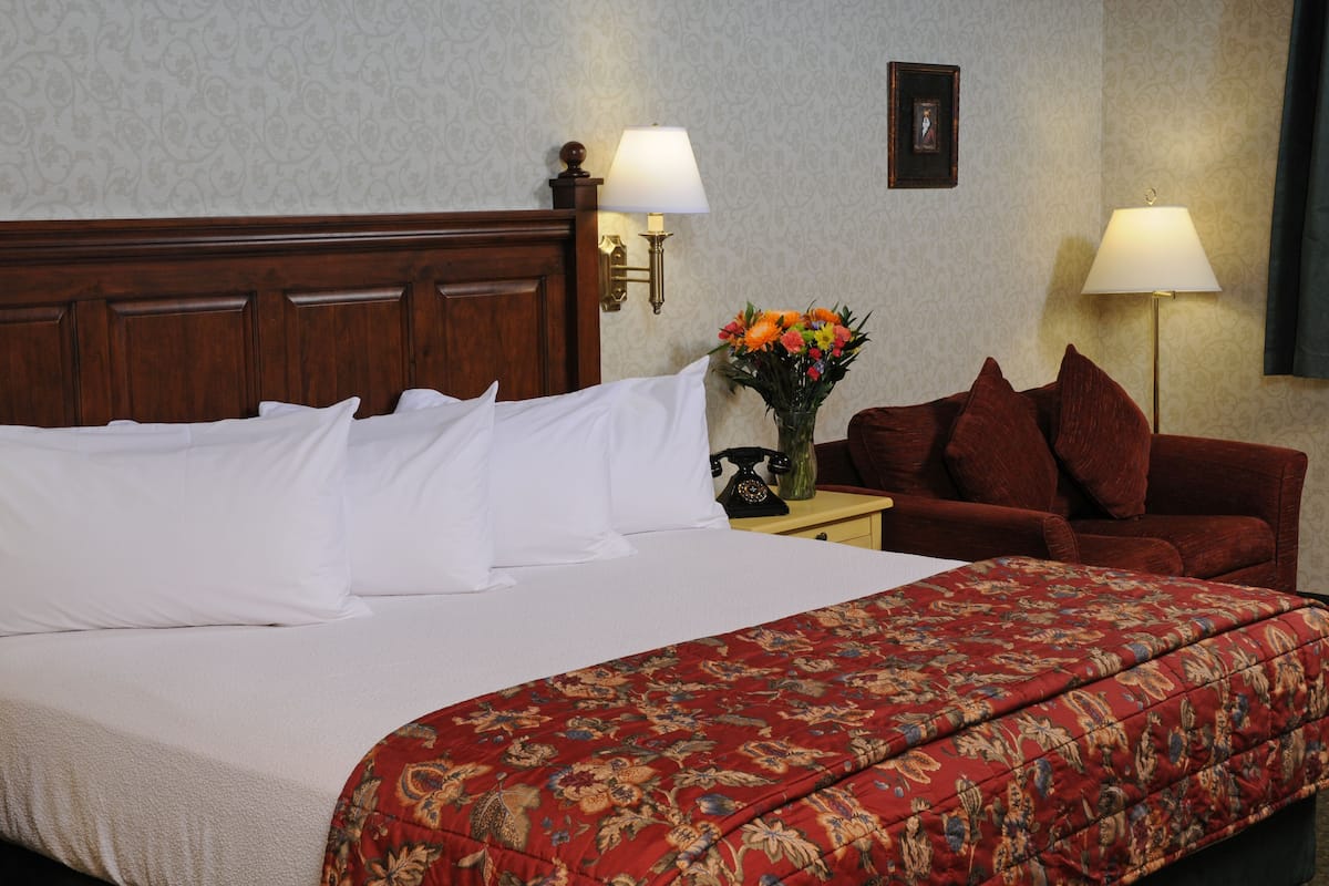 A hotel room with a large bed, a red floral bedspread, two wall-mounted lamps, a nightstand with a vase of flowers, and a red sofa.