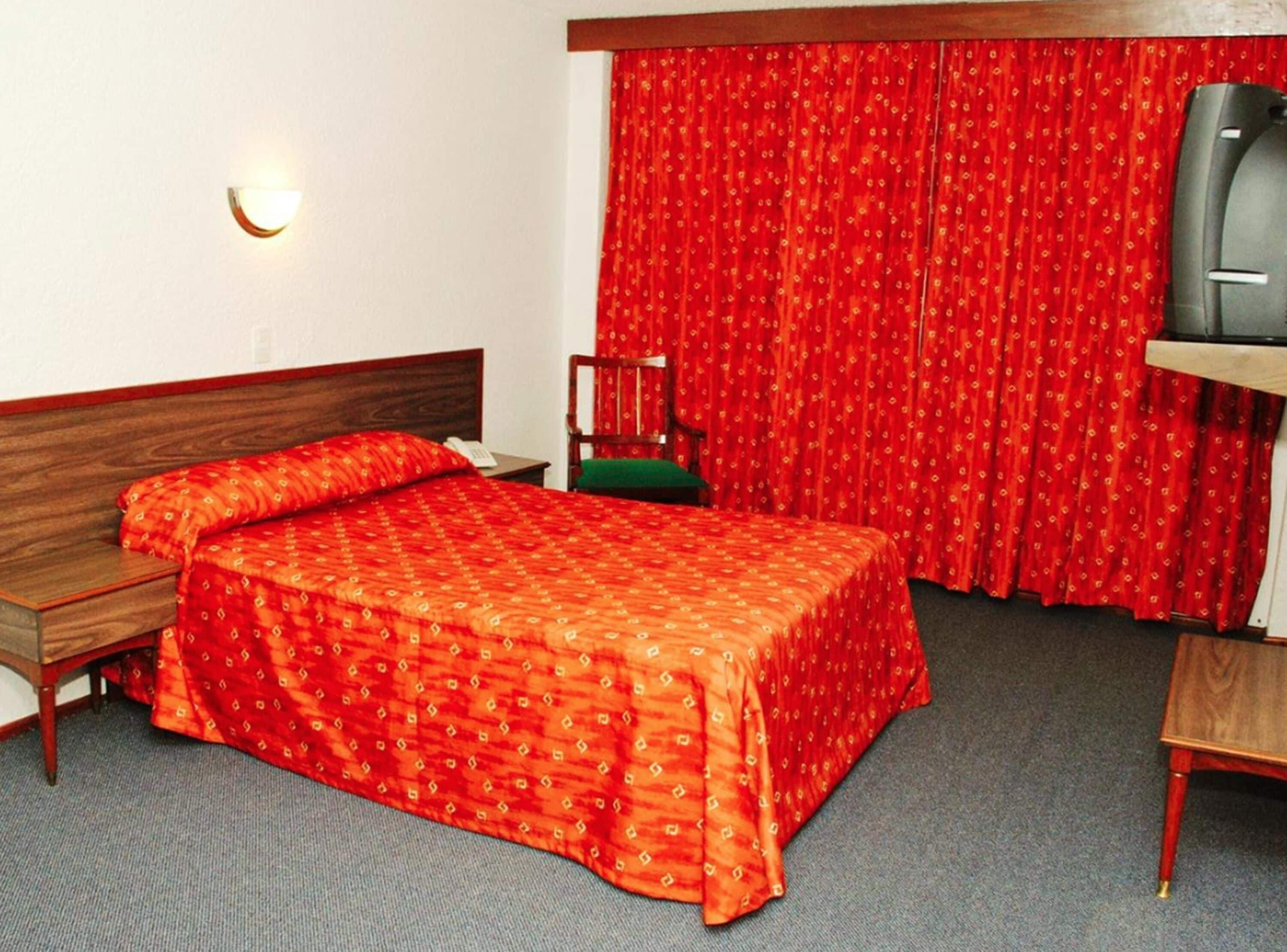 A hotel room with a wooden bed, a green chair, a desk with a television, and red curtains.