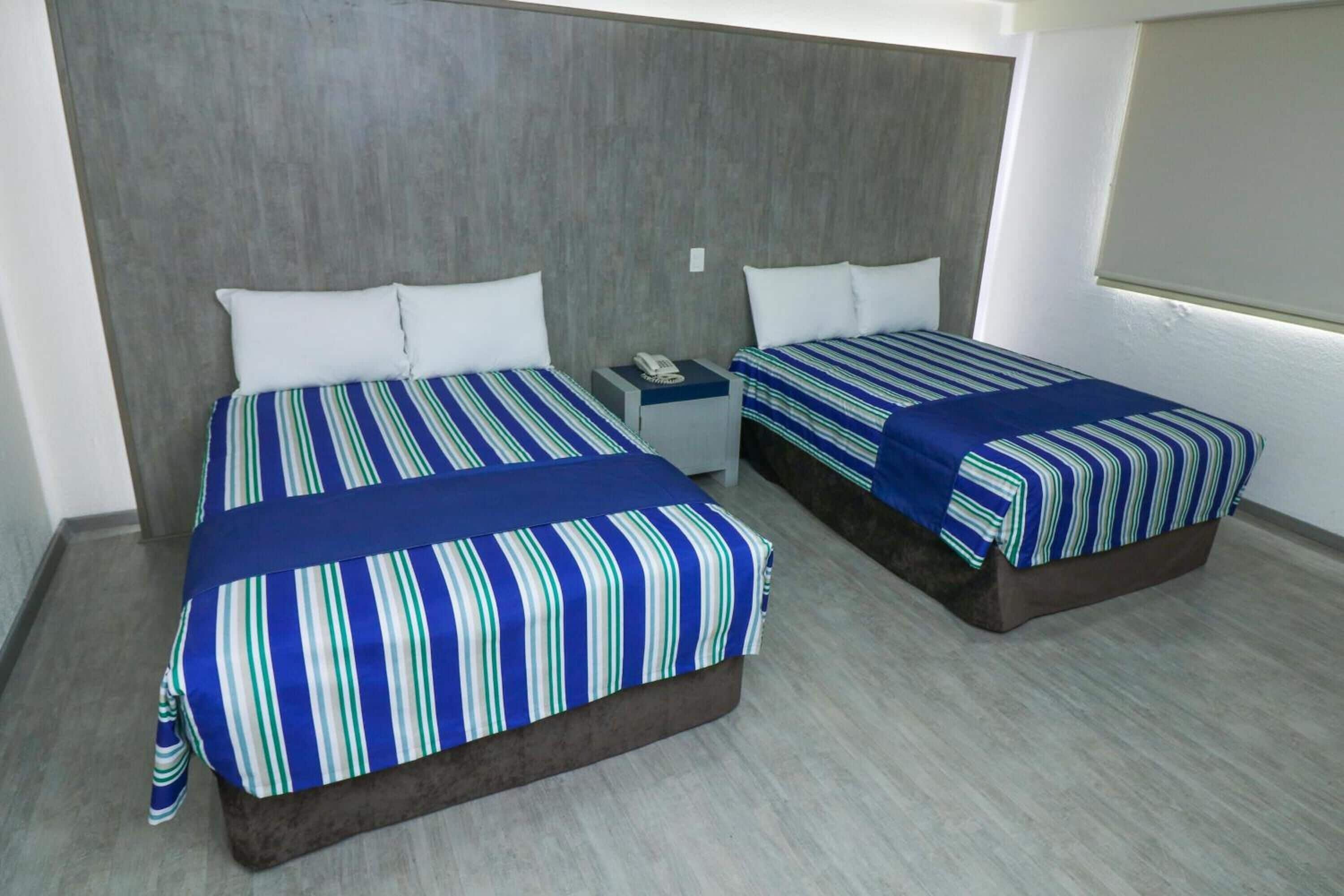 Una habitación de hotel con dos camas, cada una con una colcha a rayas y almohadas blancas. Hay una mesita de noche con un teléfono entre las camas.