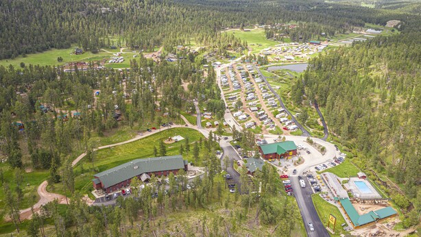 Vista aérea de un camping con varias áreas para vehículos recreativos y tiendas de campaña, un edificio central y zonas boscosas aledañas.