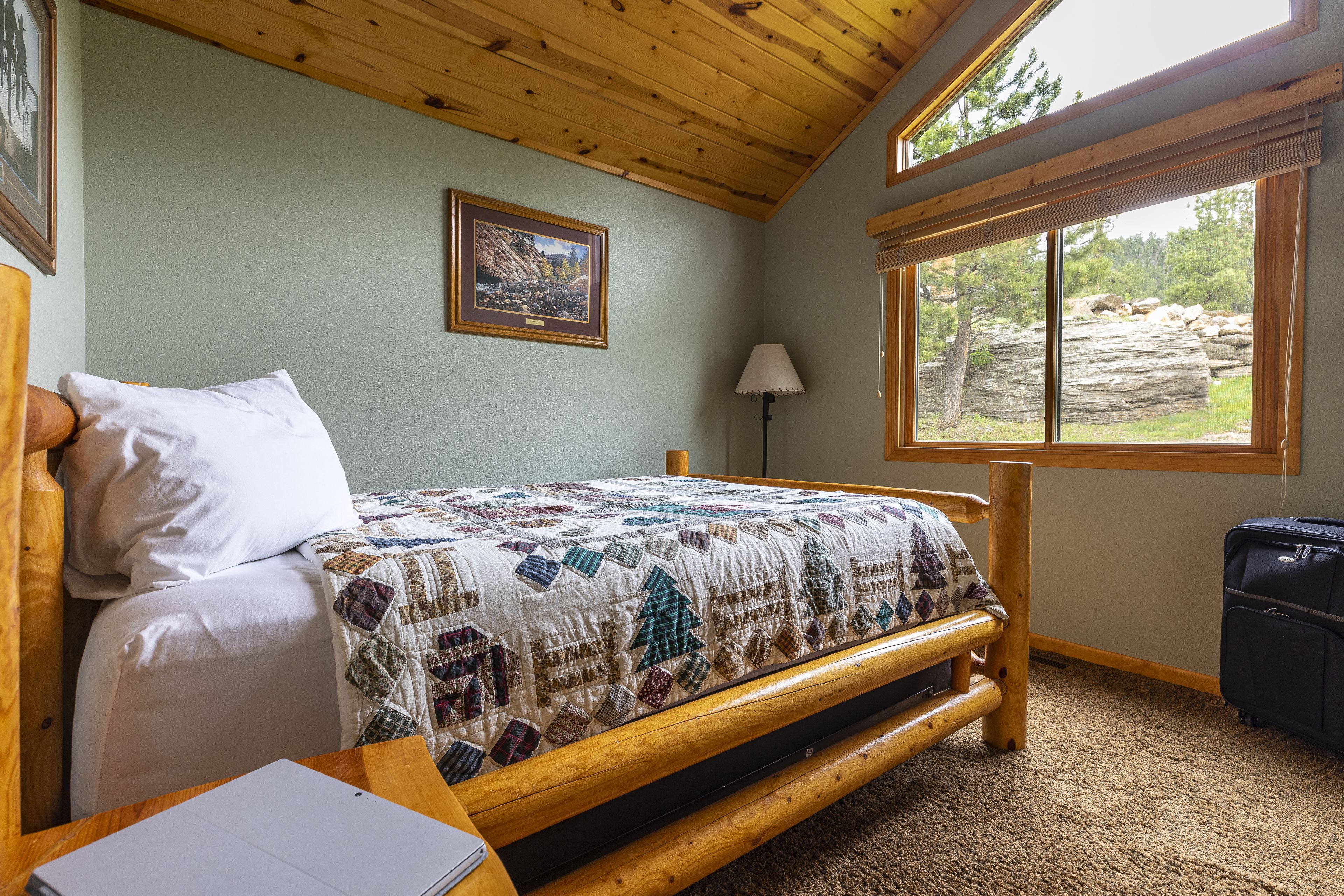 A wooden bed with a quilt, a bedside table with a notebook, a lamp, and a suitcase.