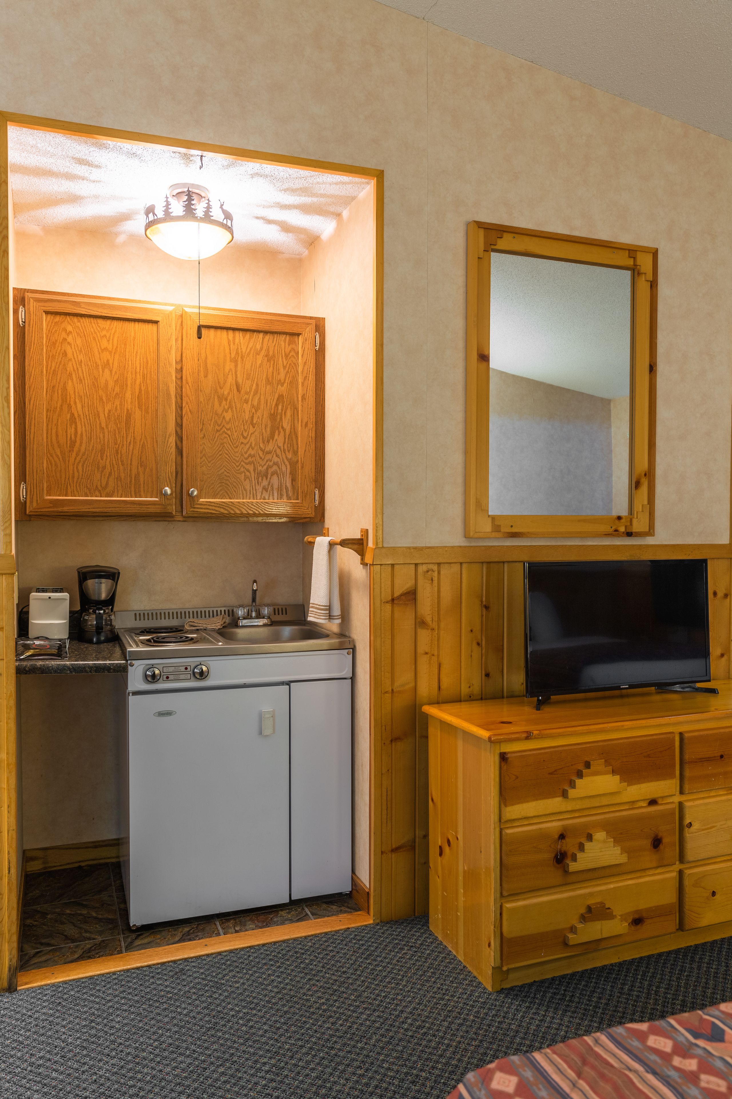 A compact kitchen with wooden cabinets, a stove, a sink, and a television on a wooden dresser.