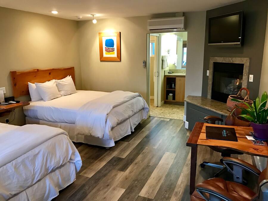 A hotel room with two beds, a desk with a laptop, a TV, a fireplace, and a bathroom visible through an open door.