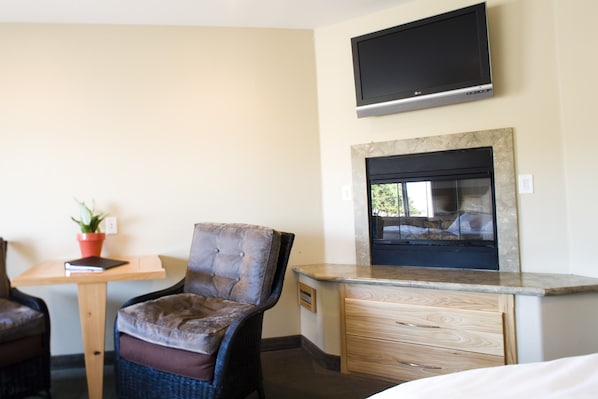 A hotel room with a flat-screen TV mounted above a fireplace, a comfortable armchair, a small table with a plant, and a wooden desk with drawers. - Cambria Shores Inn (Cambria)