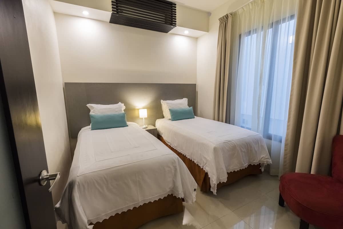 A hotel room with two beds, a red chair, a nightstand with a lamp, and a window with curtains.