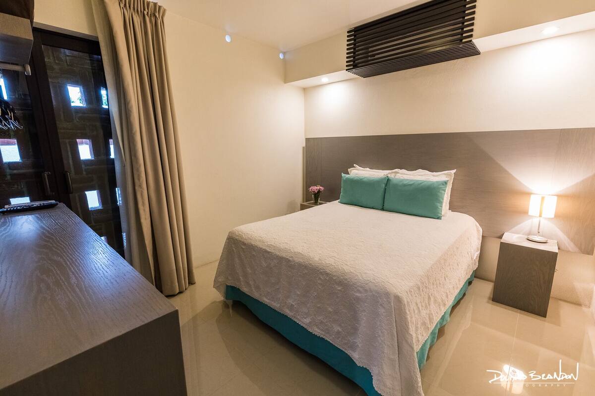 A modern hotel room with a large bed, a bedside table with a lamp, and a view through a window with curtains.