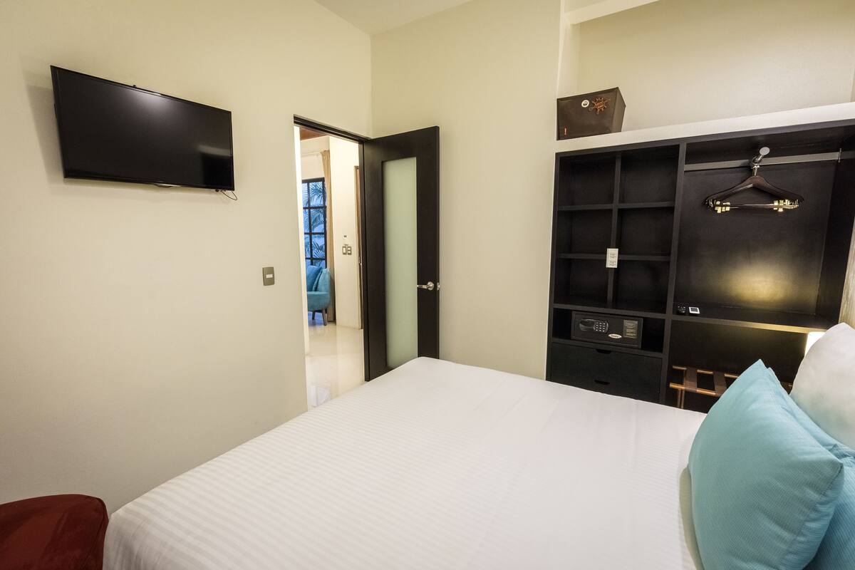 A bedroom with a bed, a TV mounted on the wall, a wardrobe, and a door leading to another room.