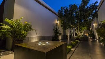 A modern courtyard with a central water feature, surrounded by potted plants and trees, and illuminated by ambient lighting.