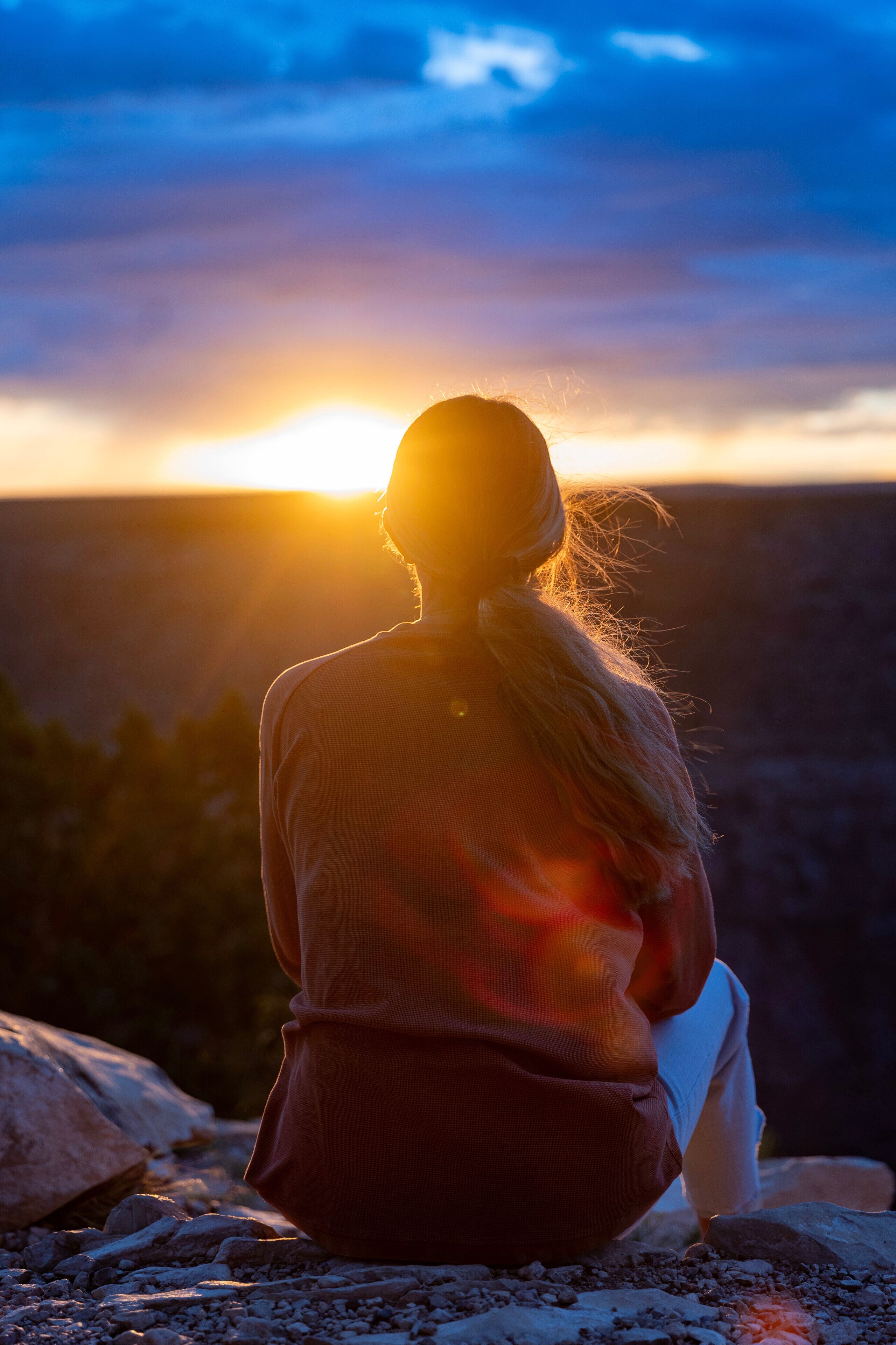 A person sitting on a rocky surface, facing away from the camera, with a sunset in the background.