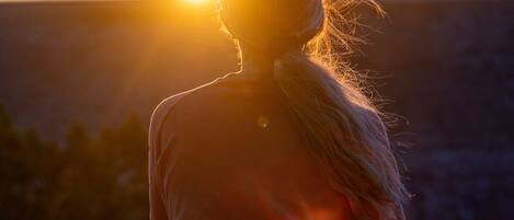 A person sitting on a rocky surface, facing away from the camera, with a sunset in the background.