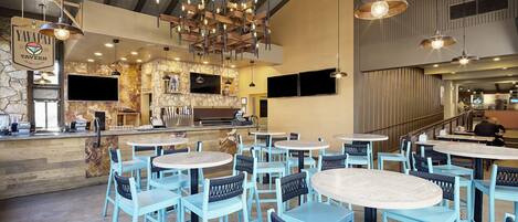 A restaurant interior with round tables and blue chairs, a stone wall, a bar area, and hanging pendant lights.