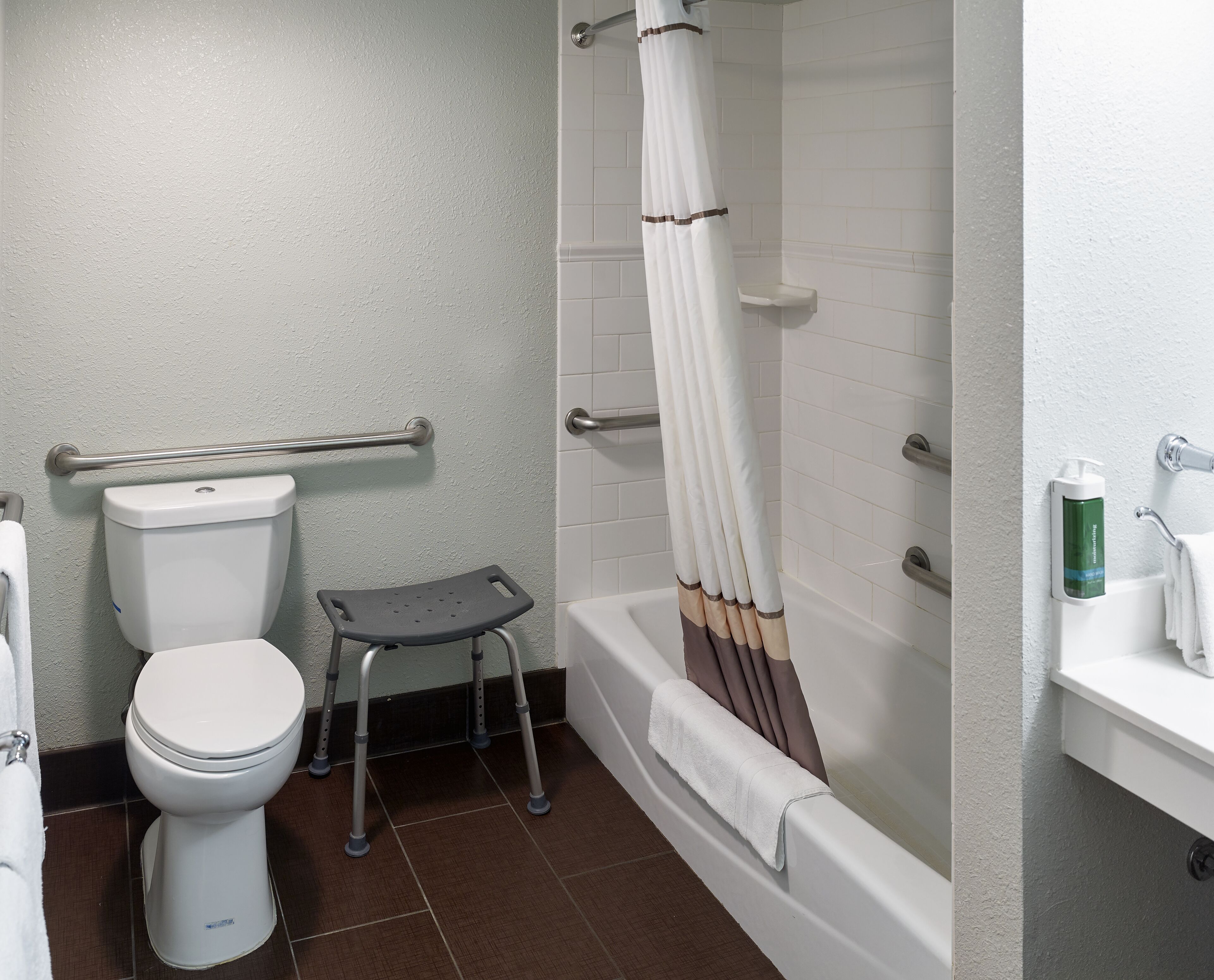 Un baño con inodoro, ducha con cortina, barra de apoyo y un pequeño taburete.