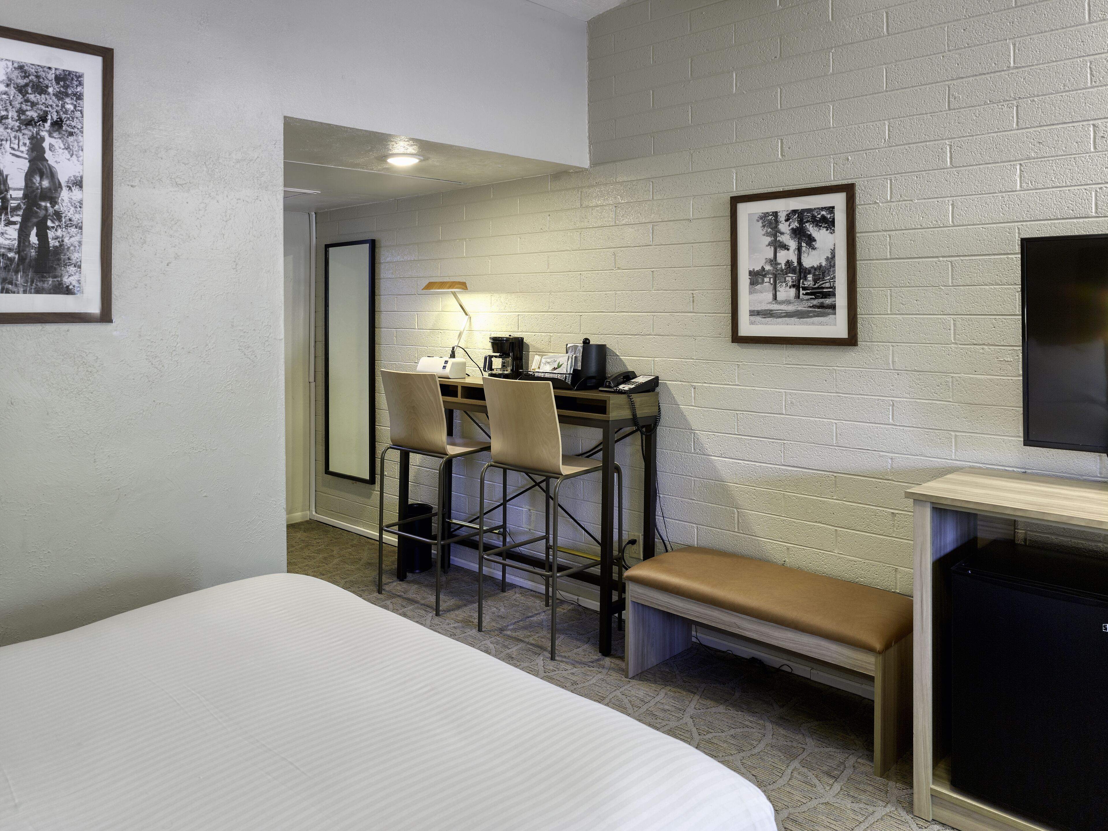 A hotel room with a bed, a desk with two chairs, a bench, a TV, and framed pictures on the walls.