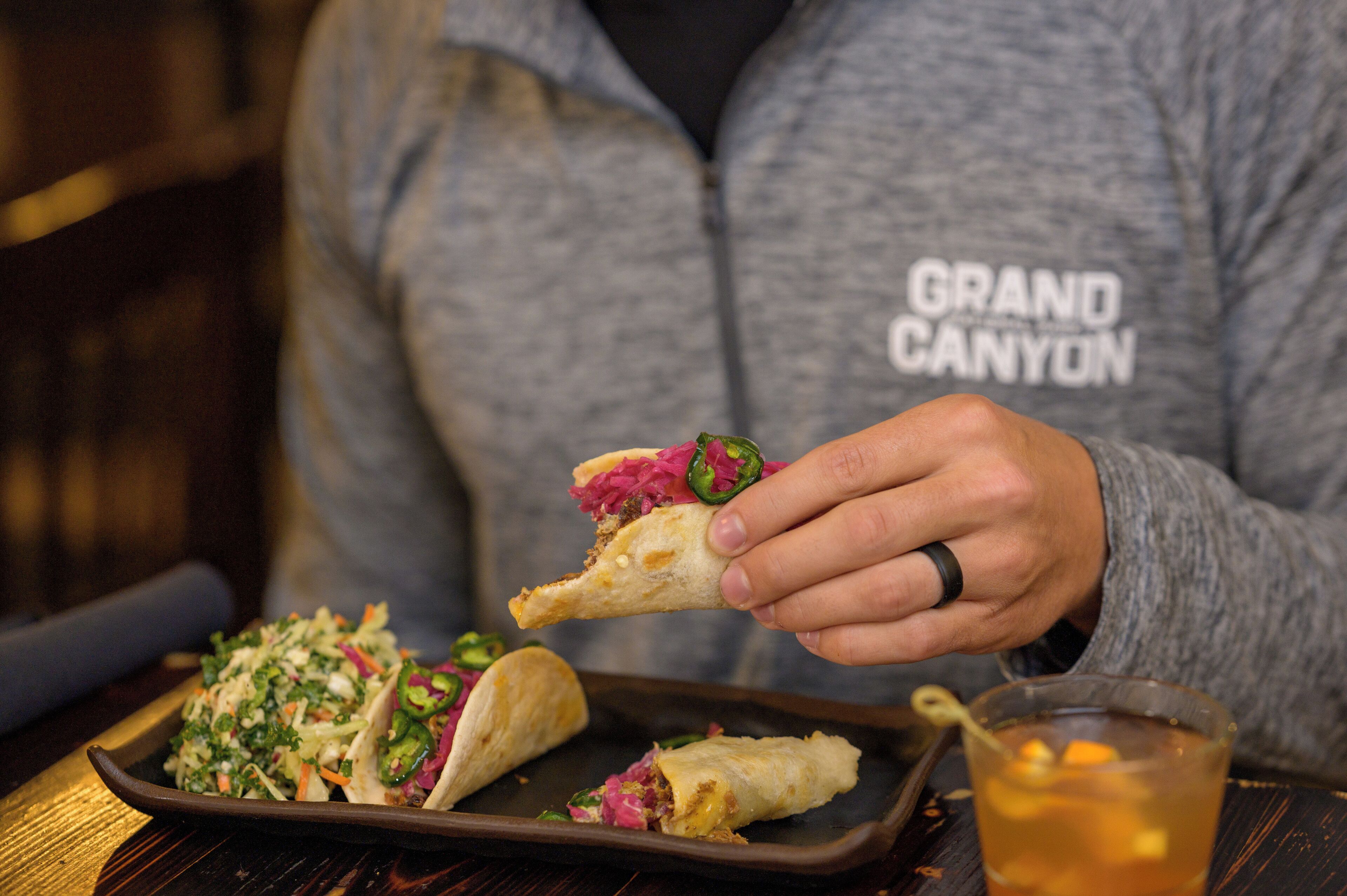 A person holding a taco with a side of coleslaw and a glass of iced tea on a wooden table.