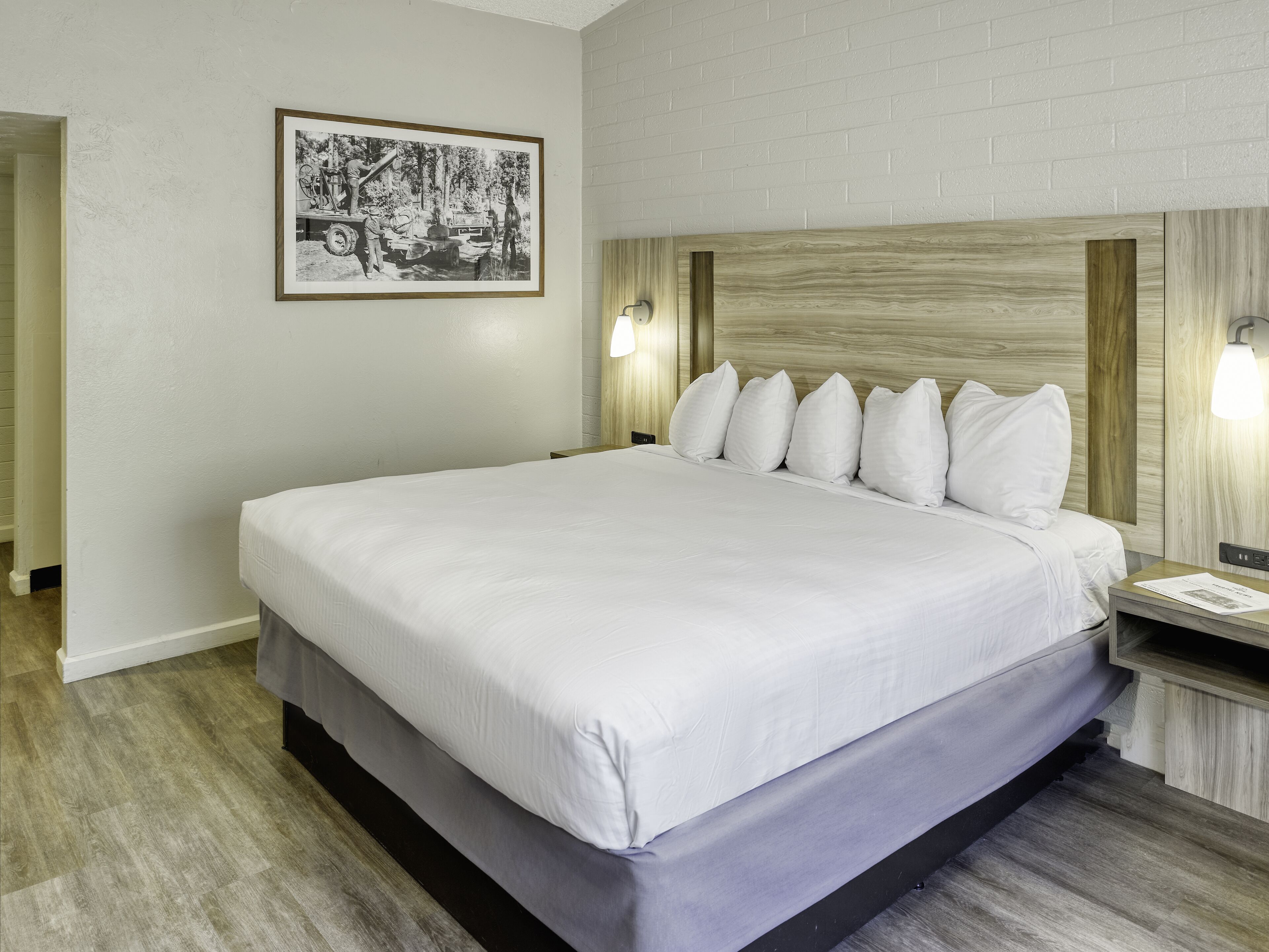 Een keurig opgemaakt bed met witte lakens, een hoofdbord met een licht houten fineer en twee bedlampjes.