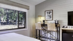 A hotel room with a bed, a desk with a lamp, chairs, a coffee maker, a refrigerator, and a large window with a view of trees.