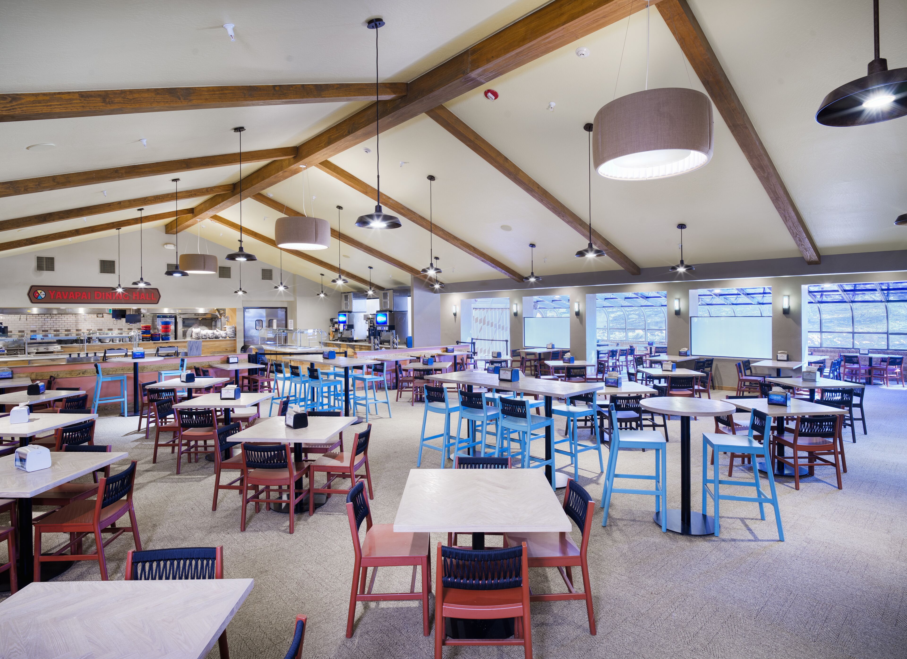 A spacious dining hall with multiple tables and chairs, high ceilings with exposed beams, and large windows providing natural light.