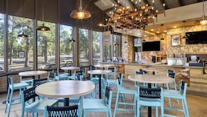A restaurant with round tables and blue chairs, large windows with a view of trees, and a stone wall with a TV.