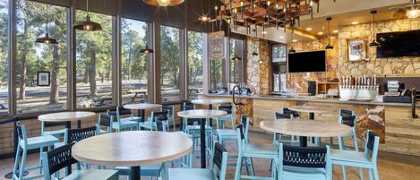 A restaurant with round tables and blue chairs, large windows with a view of trees, and a stone wall with a TV.