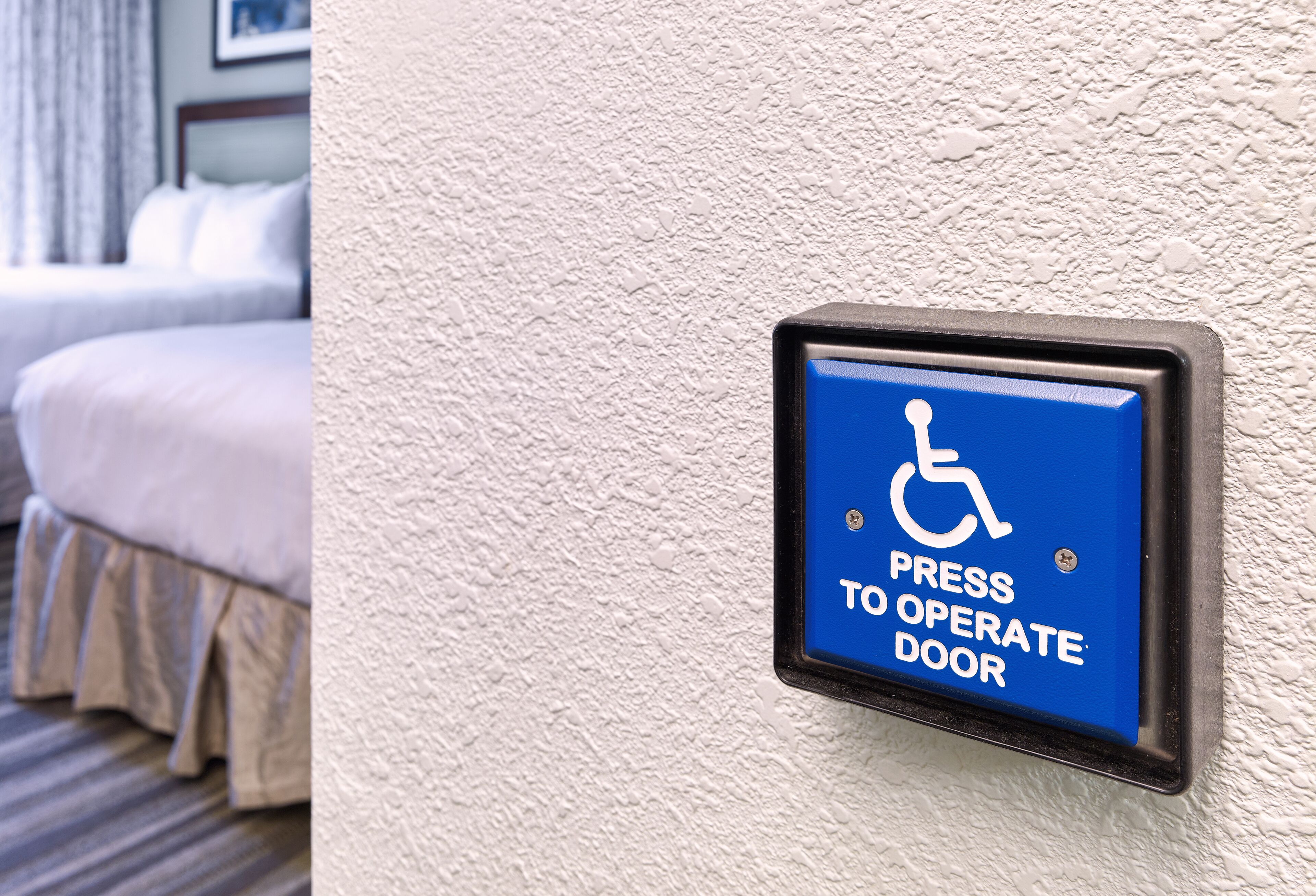 A blue sign with a wheelchair symbol and text "Press to Operate Door" on a textured wall.