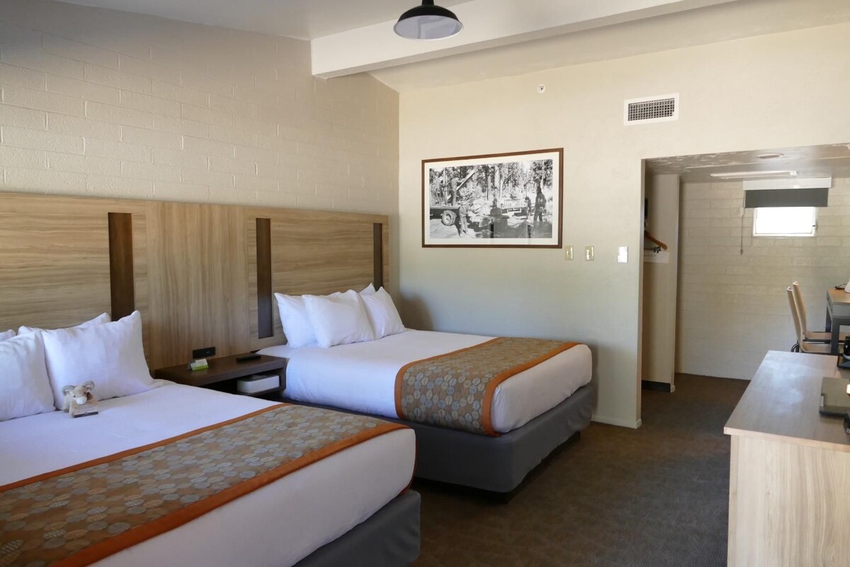 A hotel room with two beds, a desk, a chair, a framed picture on the wall, and a door leading to another room.