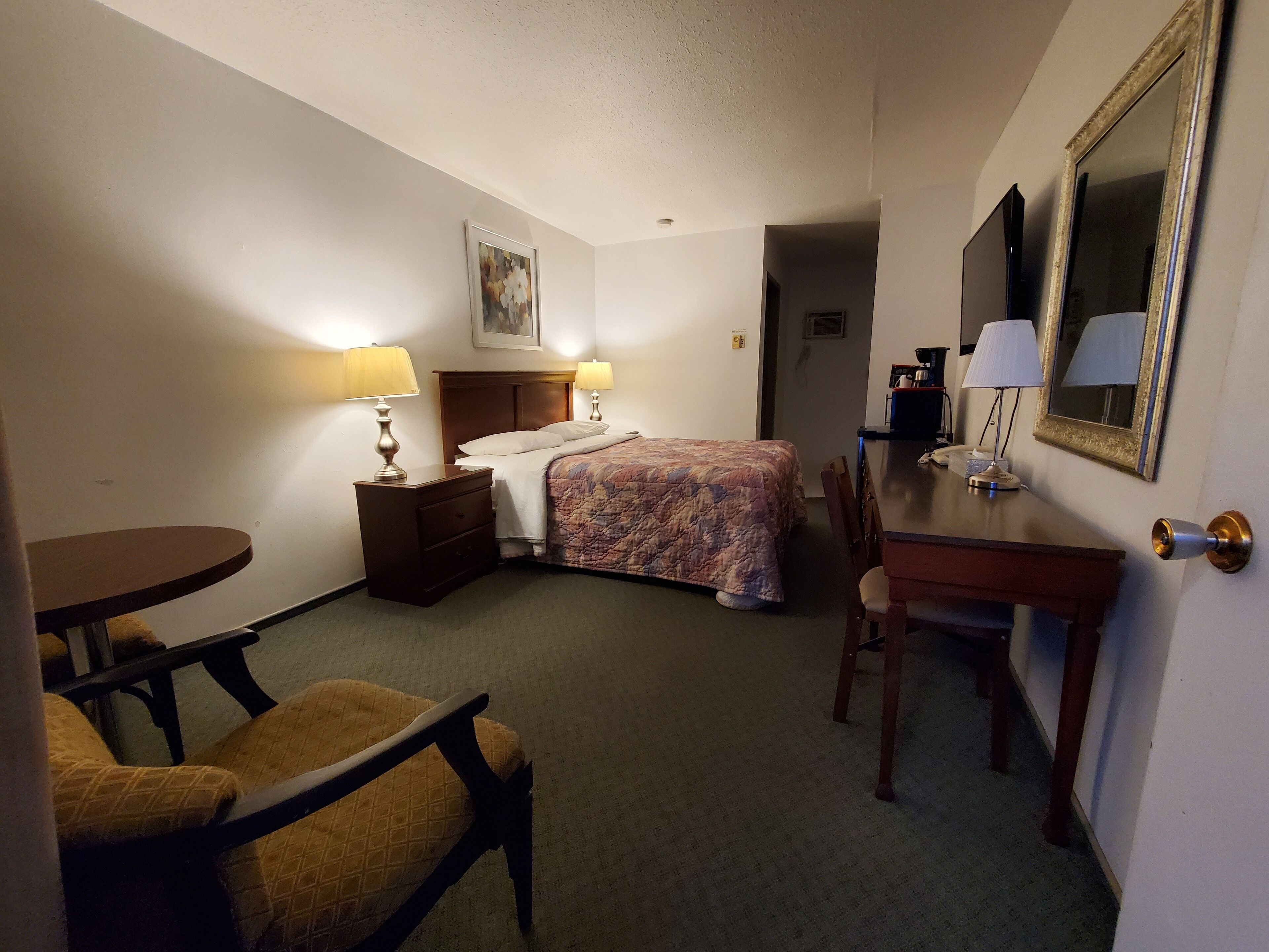 A hotel room with a bed, a desk, a chair, a table, and a mirror.