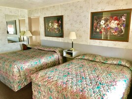 A hotel room with two beds, floral-patterned bedding, a nightstand with a lamp, a mirror, and two framed floral artworks on the wall.