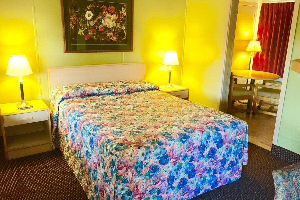 A hotel room with a floral bedspread, two bedside lamps, a nightstand, a desk with a chair, and a painting on the wall.