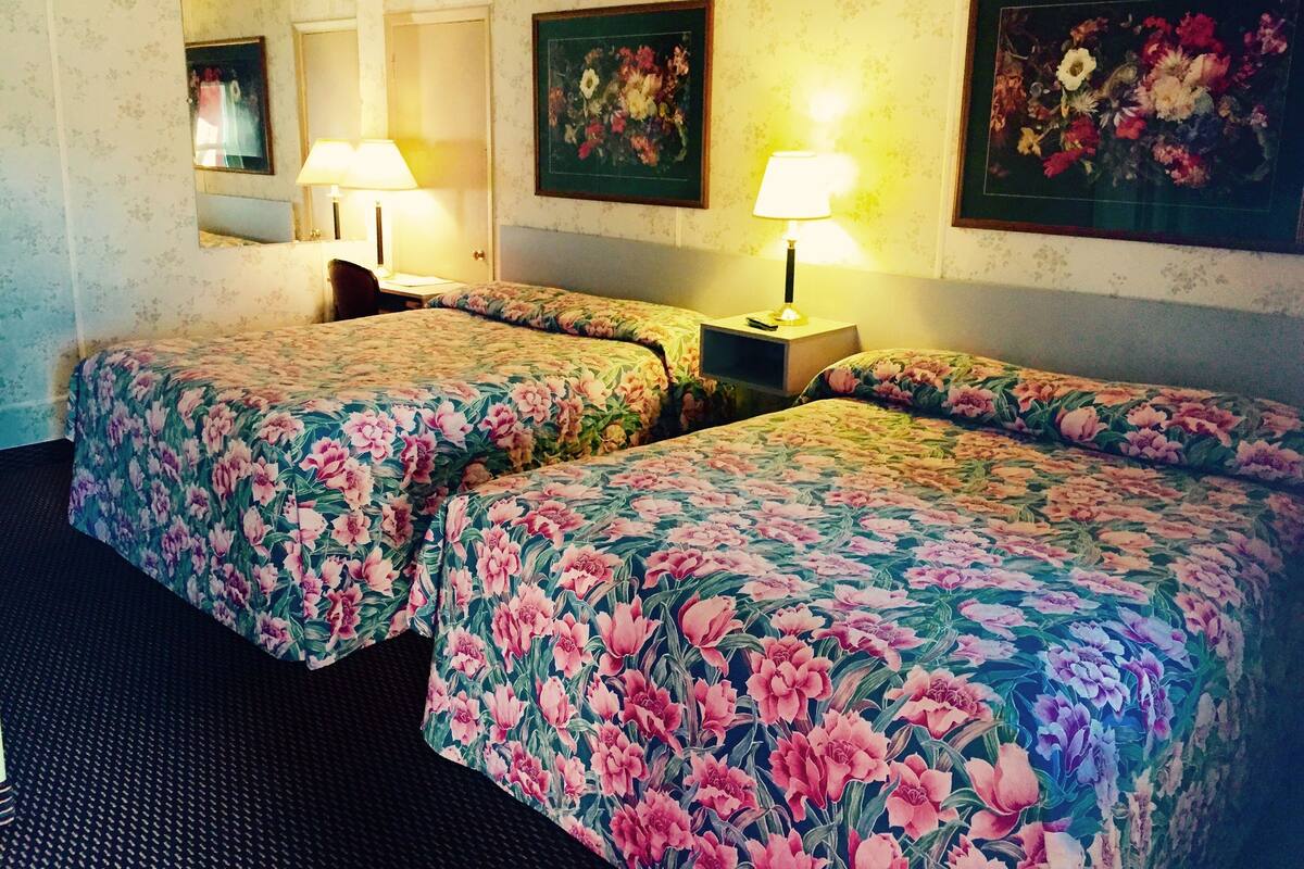 A hotel room with two beds, floral bedspreads, a nightstand with a lamp, a mirror, and a framed floral painting on the wall.
