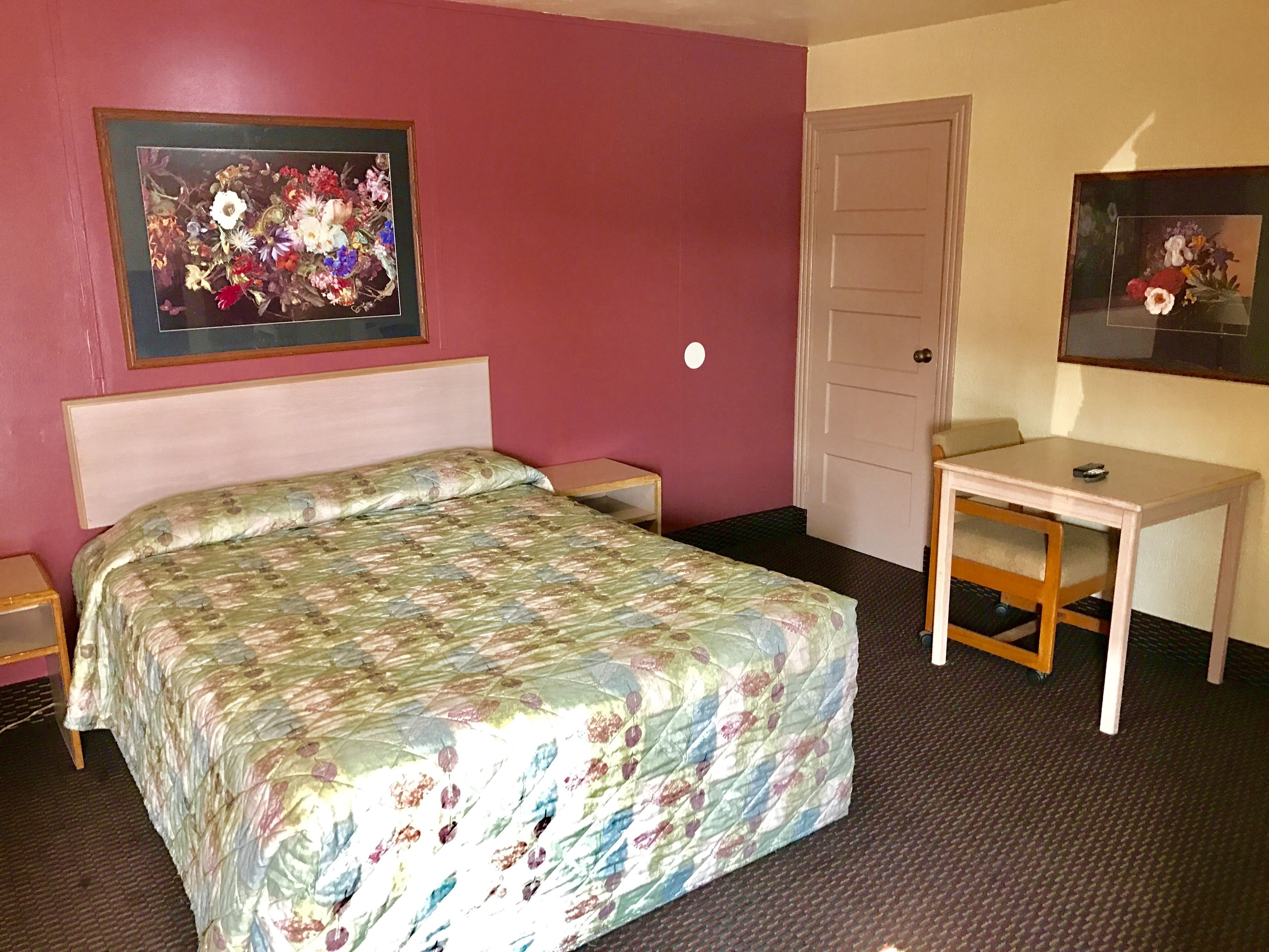 A hotel room with a bed, a desk, and two framed floral paintings on the walls.