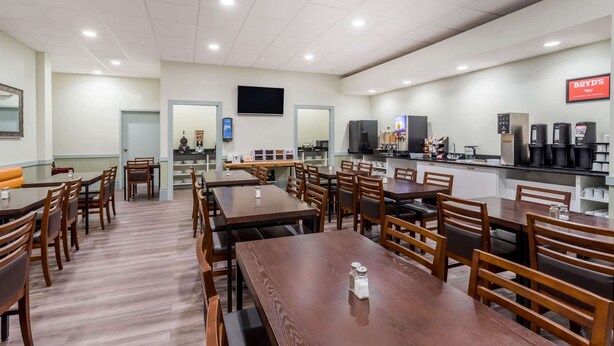 Un comedor amplio con varias mesas y sillas de madera, una zona de cocina con máquinas de café y un televisor de pantalla plana montado en la pared.