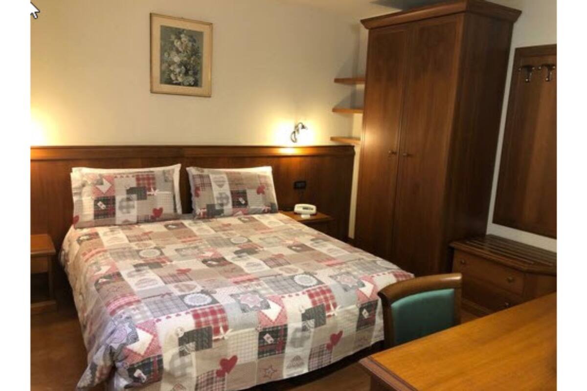 A bedroom with a large bed covered in a quilt, a wooden wardrobe, a nightstand with a lamp, and a framed picture on the wall.