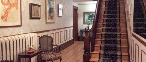 A grand staircase with a dark patterned carpet, a radiator, and framed artwork on the walls.