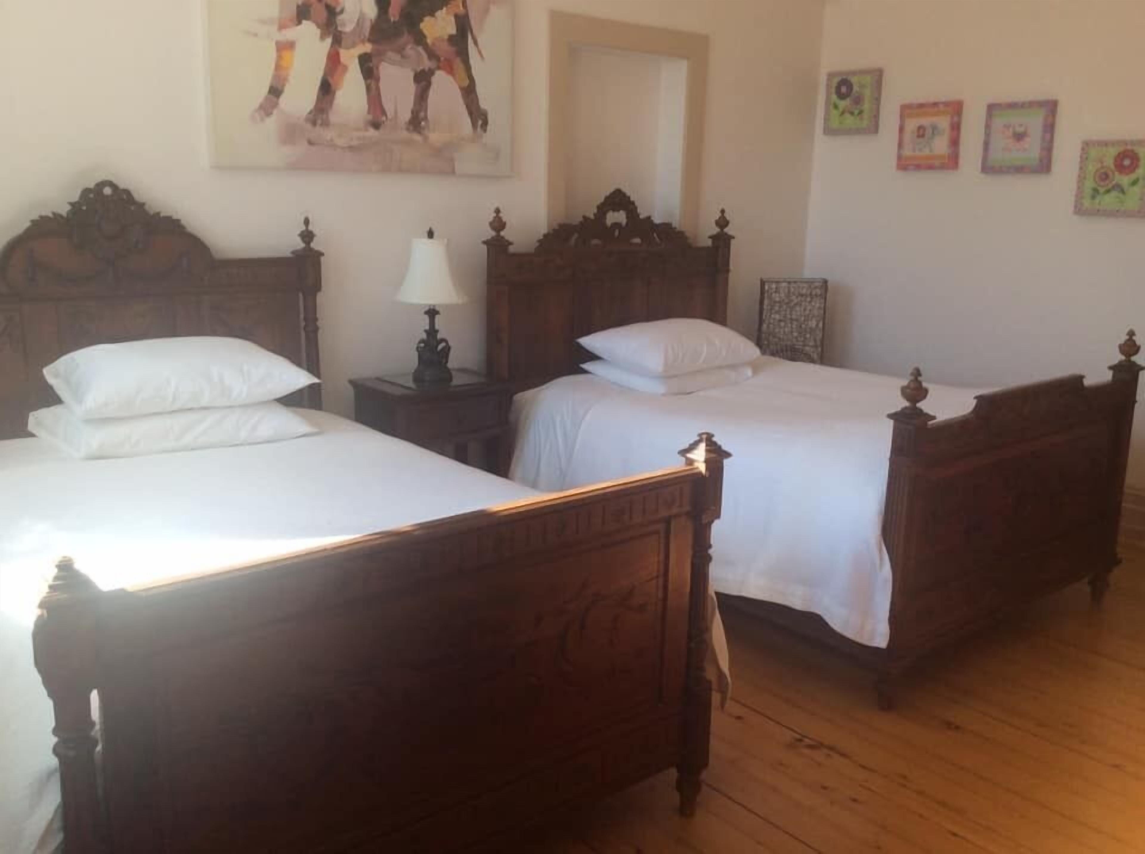 Una habitación con dos camas de madera, ropa de cama blanca, una mesita de noche de madera con una lámpara y un cuadro en la pared.