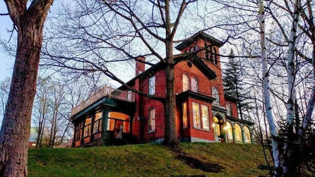 Una casa grande de ladrillo rojo con varias ventanas y un balcón, rodeada de árboles.