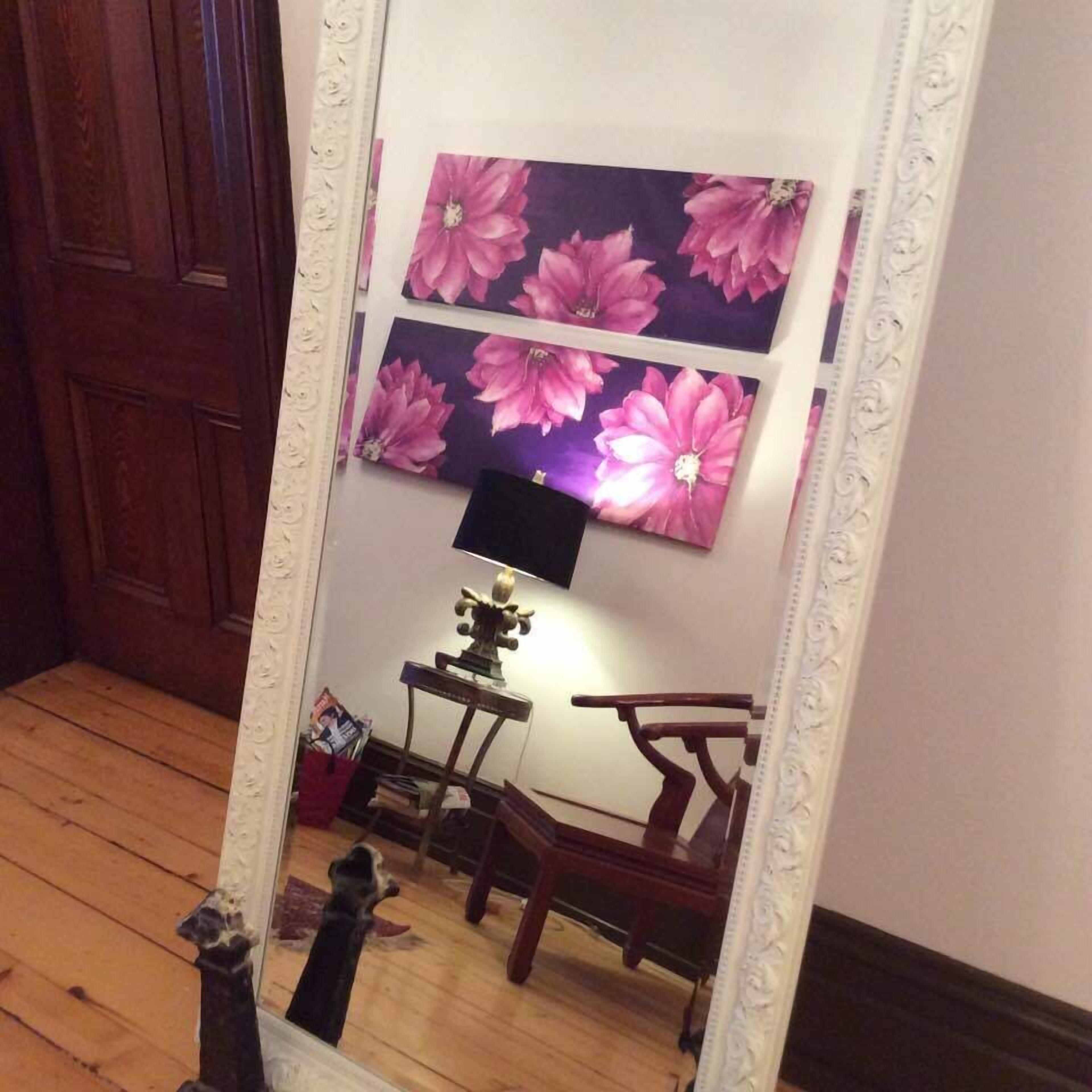 Une pièce avec un plancher en bois, un miroir qui reflète un mur orné de deux grandes peintures florales, une petite table avec une lampe et une chaise.
