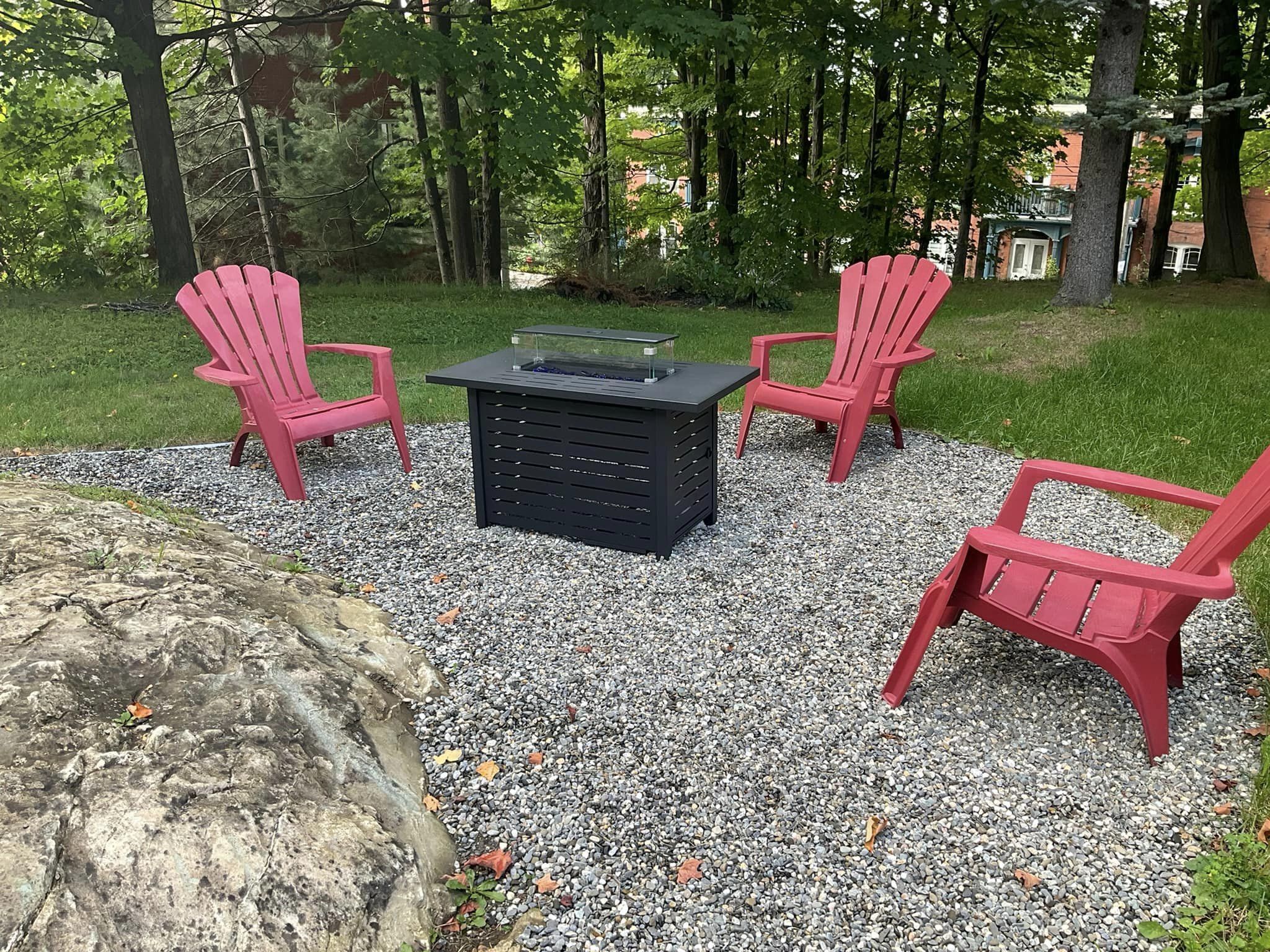 An outdoor seating area with four red Adirondack chairs and a black table with a glass top, surrounded by gravel and trees.