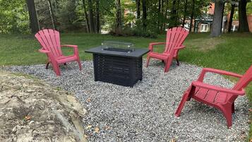 An outdoor seating area with four red Adirondack chairs and a black table with a glass top, surrounded by gravel and trees.