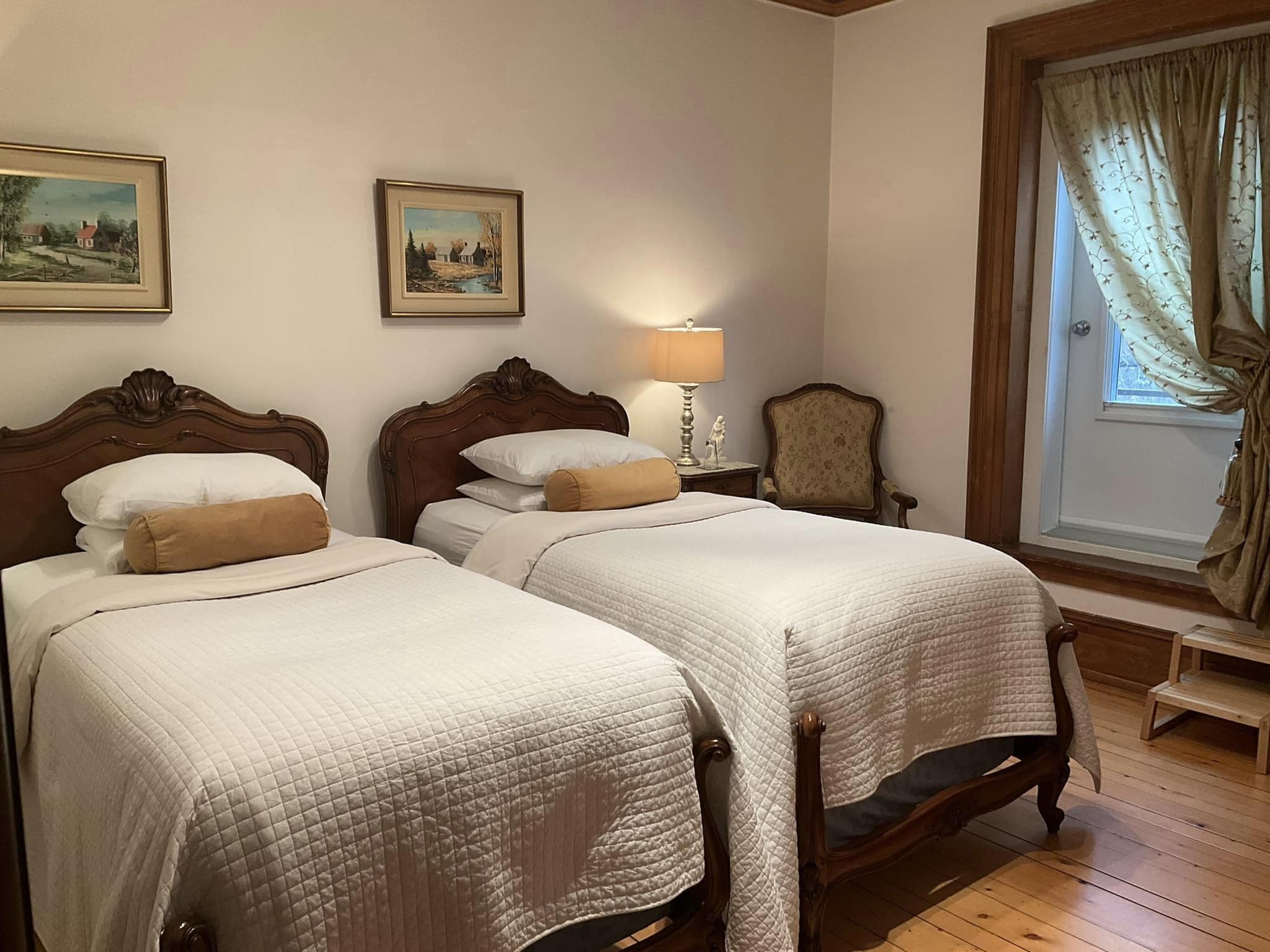 A room with two beds, wooden headboards, a nightstand with a lamp, a chair, and two framed pictures on the wall.