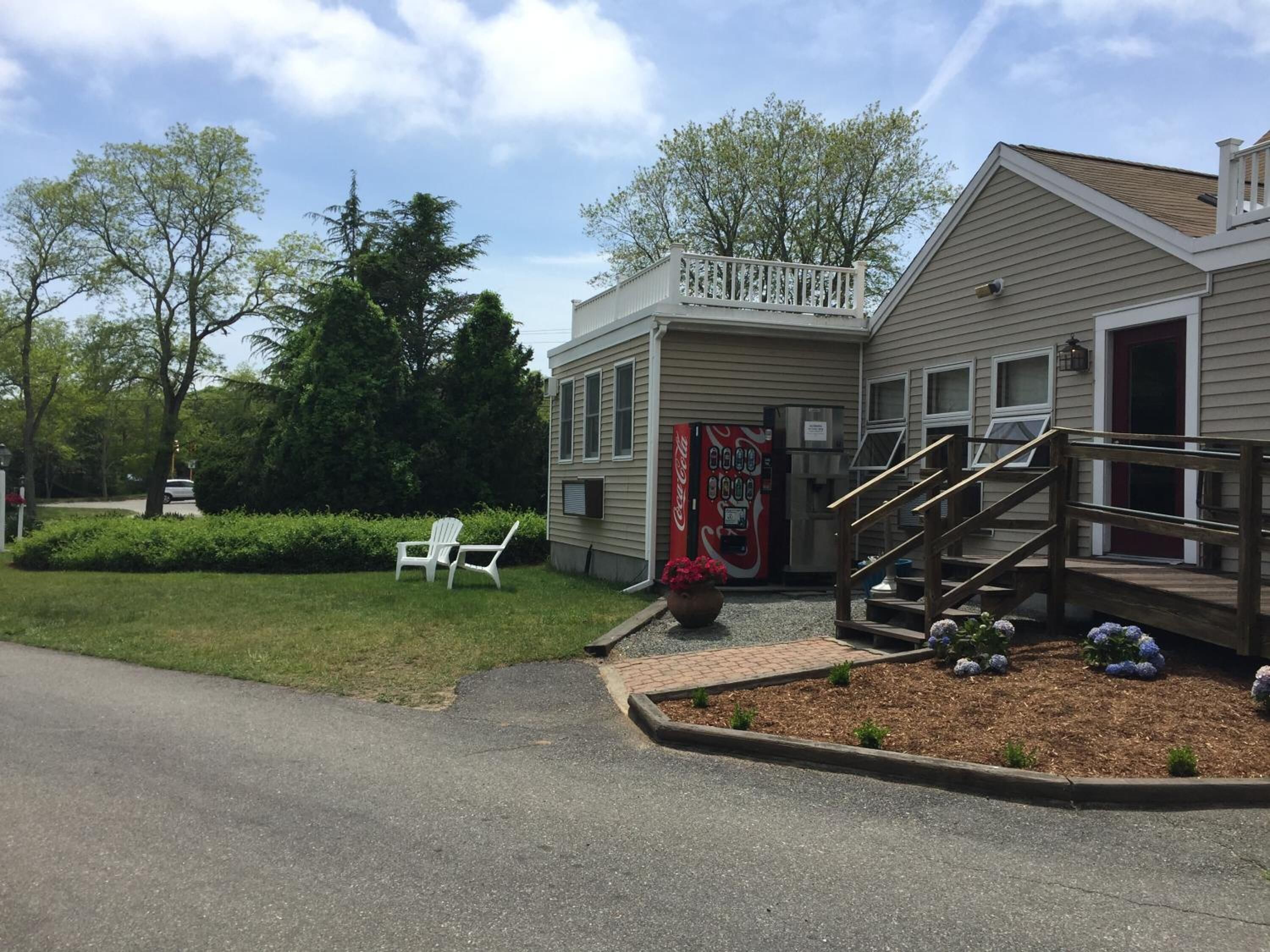 A two-story building with a front porch, a Coca-Cola vending machine, and a small garden area with flowers and shrubs.